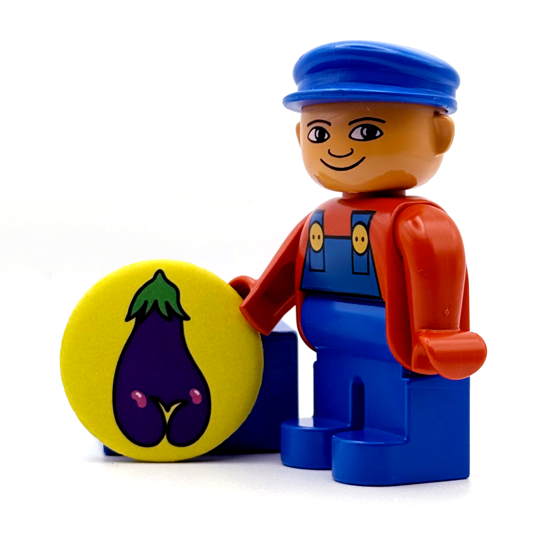 Toy figure with a blue hat and overalls holding a yellow magnet with an eggplant design. The eggplant has an asscrack.