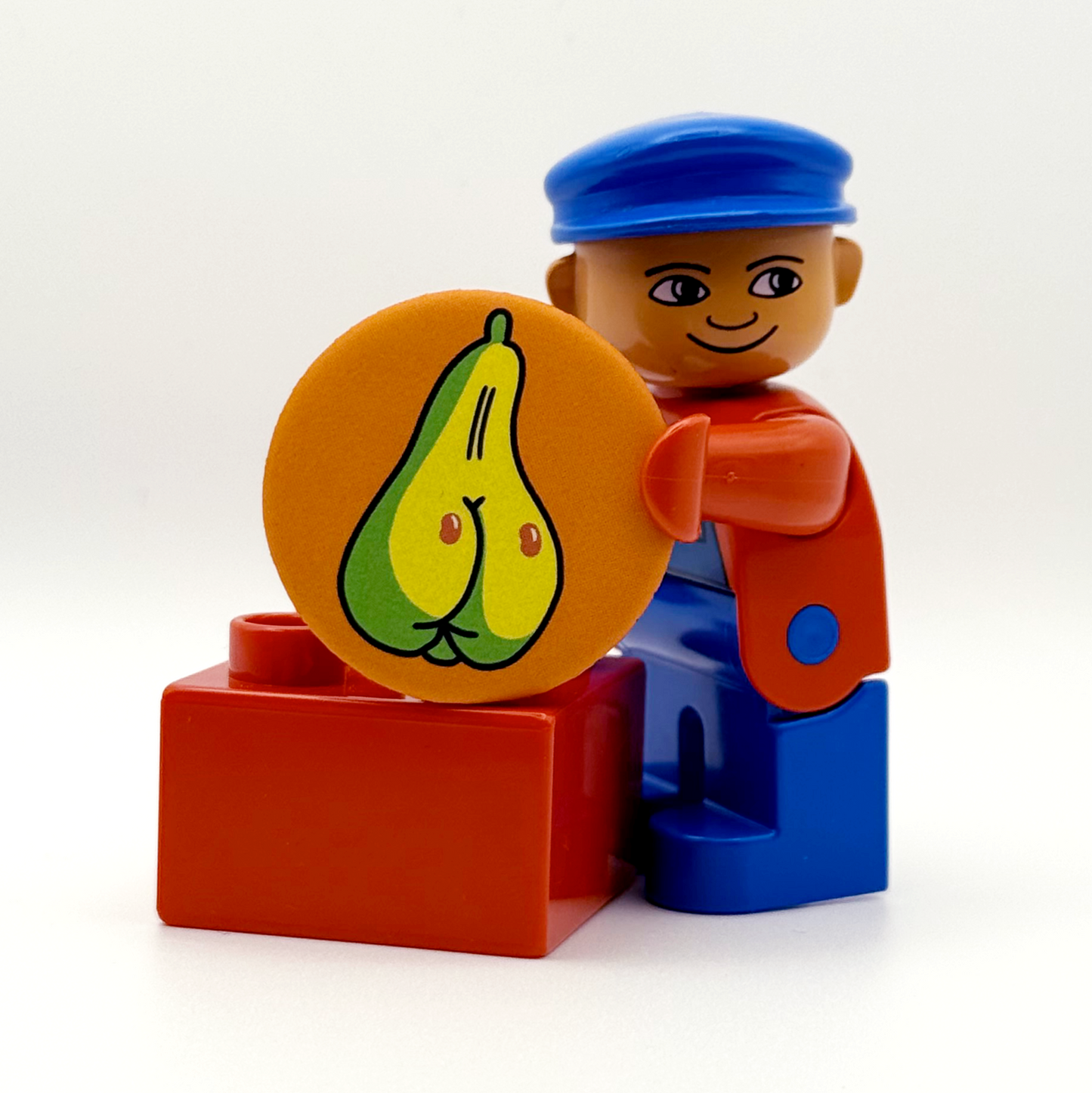 LEGO figure with a blue hat and orange shirt holding a round magnet with a pear on a white background. The pear has an asscrack.