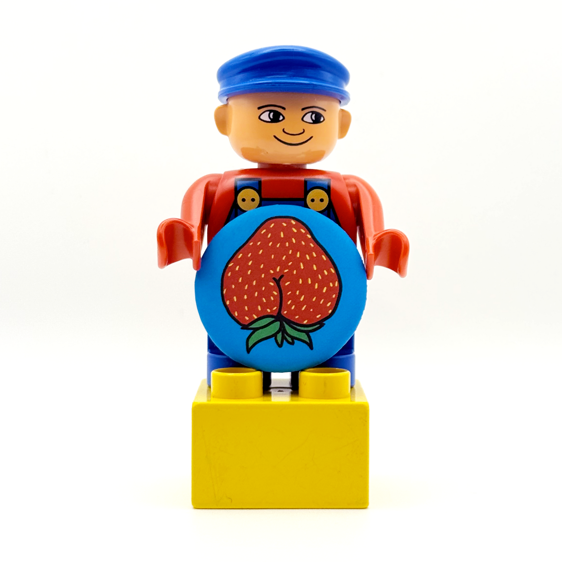 It's a Duplo worker toy holding a fruity and fancy tiny ass magnet. The magnet features a strawberry with an asscrack with a matte finish. 